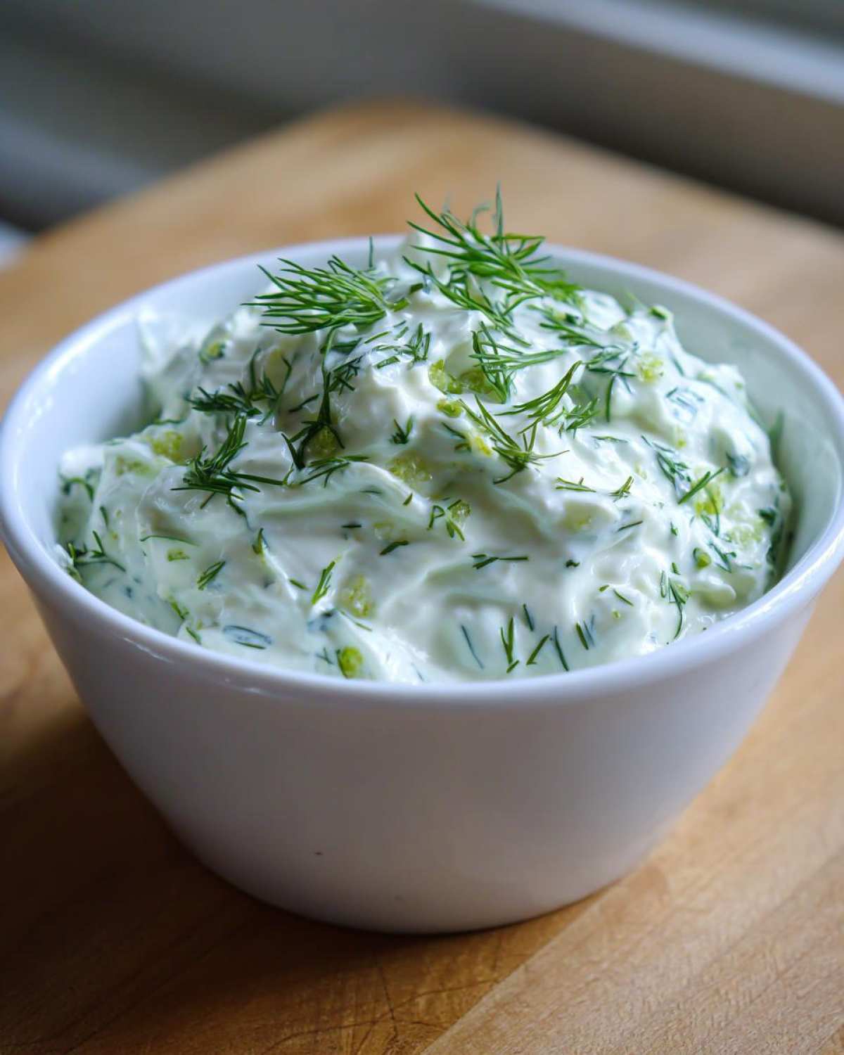 A white bowl filled with creamy Homemade Tzatziki Sauce, garnished generously with fresh dill.