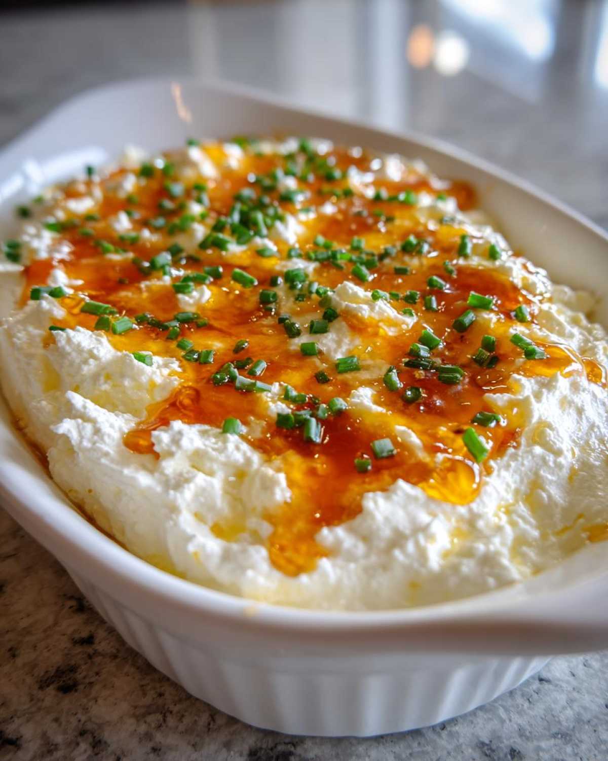 A white dish filled with creamy Hot Honey Ricotta Dip, drizzled with honey and topped with fresh green chives.