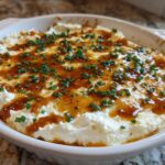 A round white baking dish filled with creamy Hot Honey Ricotta Dip, drizzled with honey and topped with fresh chives.