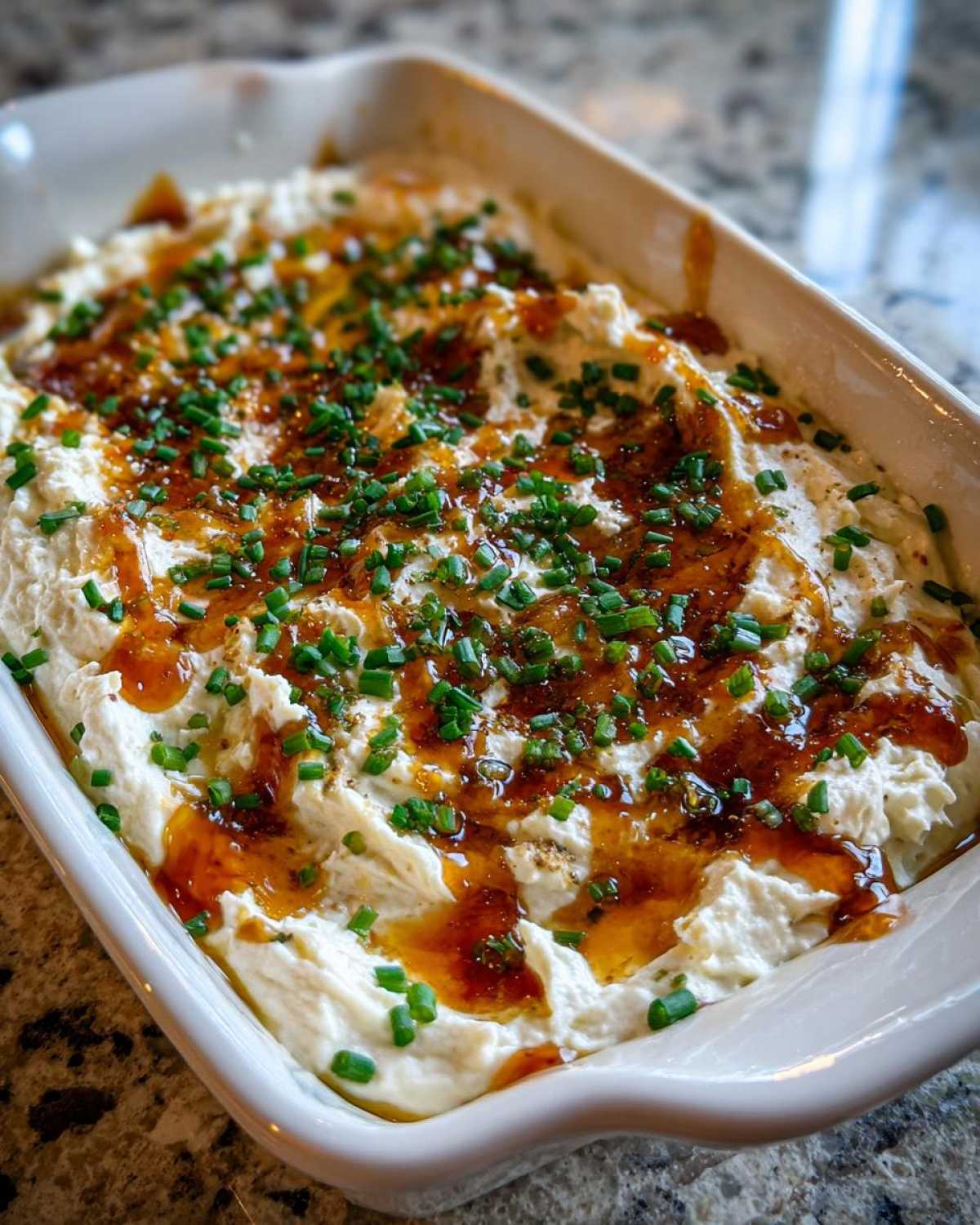 A white baking dish filled with creamy Hot Honey Ricotta Dip, drizzled with honey and topped with fresh chives.