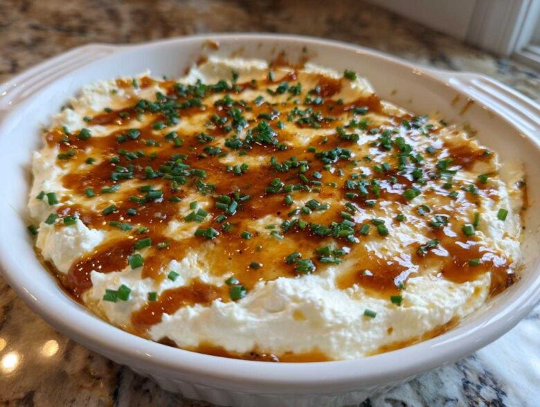 A round white baking dish filled with creamy Hot Honey Ricotta Dip, drizzled with honey and topped with fresh chives.
