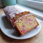 A loaf of moist Rhubarb Bread, partially sliced, topped with a thick white glaze, showing pink rhubarb pieces inside.