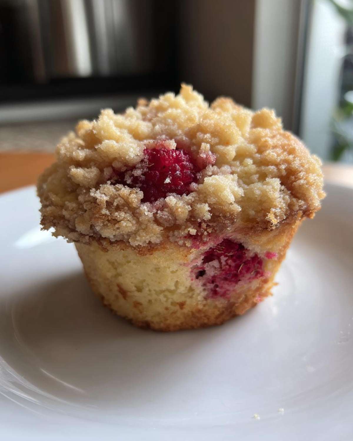 A single, golden Raspberry Streusel Muffins sits on a white plate, featuring a thick, sugary crumb topping and visible bright red raspberries.